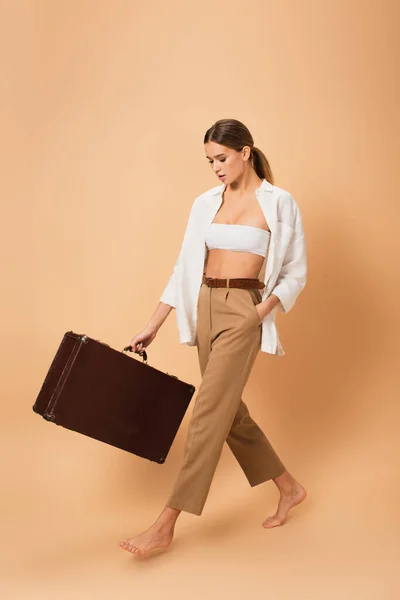 Jeune femme en pantalon et chemise déboutonnée marchant pieds nus avec valise vintage sur fond beige — Photo de stock