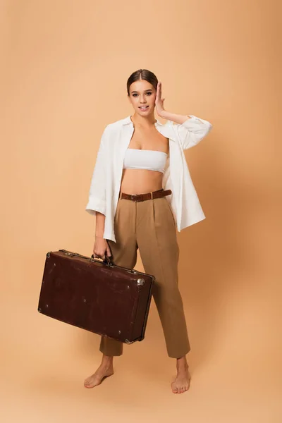 Barefoot, stylish woman with retro suitcase touching face while standing on beige background — Stock Photo