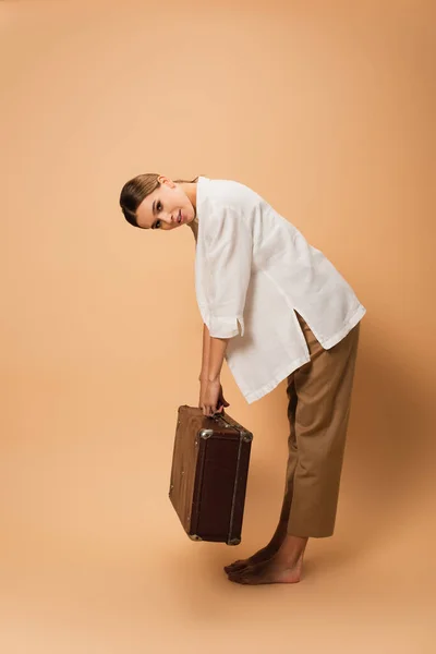 Barefoot woman in trousers and white shirt bending with retro suitcase on beige background — Stock Photo