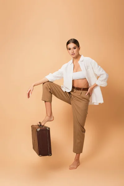 Barefoot woman in stylish clothes stepping on retro suitcase on beige background — Stock Photo