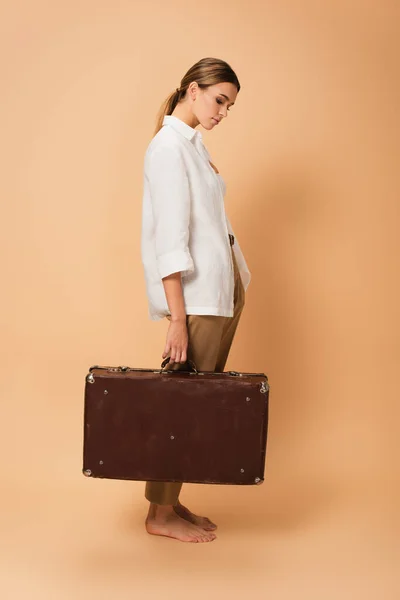 Side view of barefoot woman in white shirt and trousers holding vintage suitcase on beige background — Stock Photo