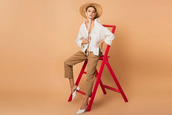 Sensual woman sitting on ladder in unbuttoned shirt, pants and straw hat on beige background — Stock Photo