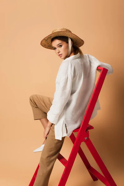 Donna alla moda in camicia bianca, pantaloni e cappello di paglia guardando la fotocamera su scala rossa su sfondo beige — Foto stock