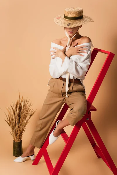 Mujer sensual en camisa, pantalones y sombrero de paja abrazándose mientras está sentada en la escalera roja en beige - foto de stock