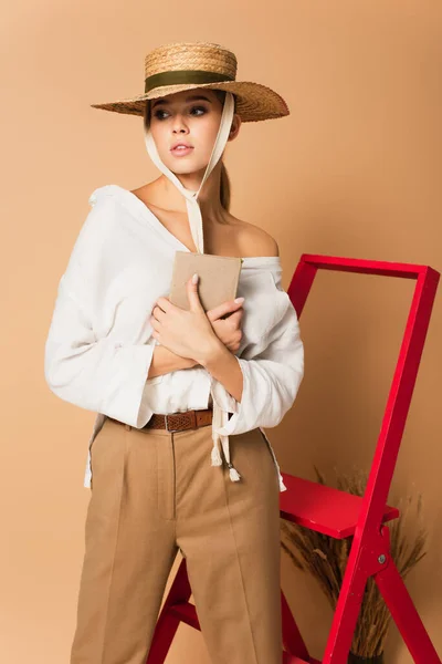 Young woman in white shirt, trousers and straw hat holding book on beige background — Stock Photo