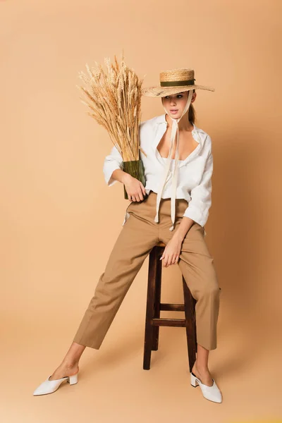 Fashionable woman in straw hat sitting on wooden stool with vase of spikelets on beige — Stock Photo
