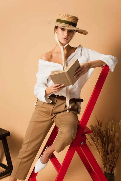 Молода жінка в модному одязі і солом'яному капелюсі позує з оголеним плечем і книгою на бежевому — стокове фото