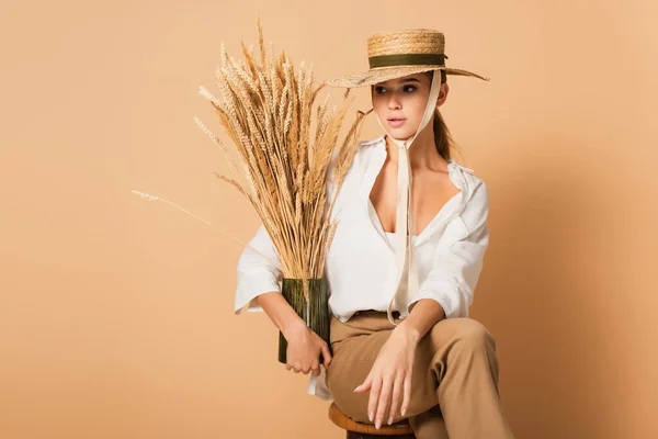 Woman in white shirt and straw hat holding vase with spikelets on beige background — Stock Photo