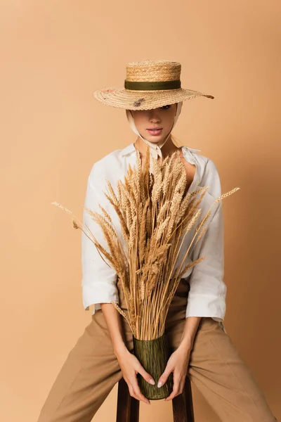 Sensual woman in straw hat and white shirt holding vase of wheat spikelets and looking at camera on beige — Stock Photo