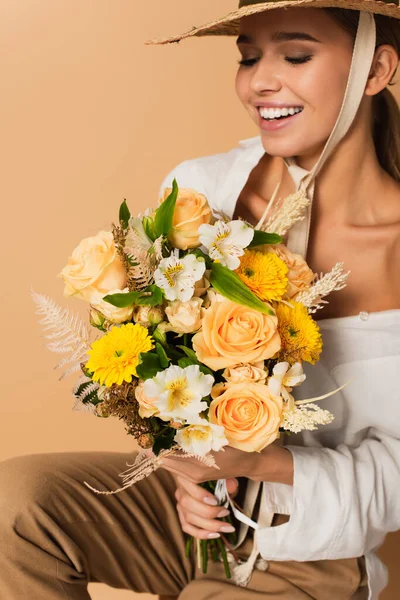 Giovane donna felice in cappello di paglia che tiene bouquet di fiori diversi isolati su beige — Foto stock