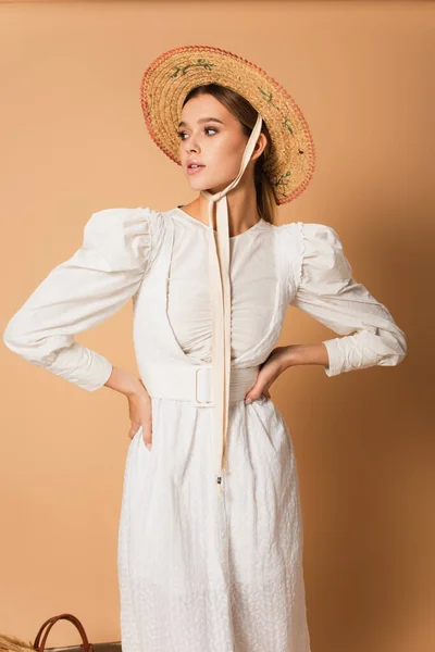 Giovane donna in abito bianco e cappello di paglia in piedi con le mani sui fianchi su beige — Foto stock