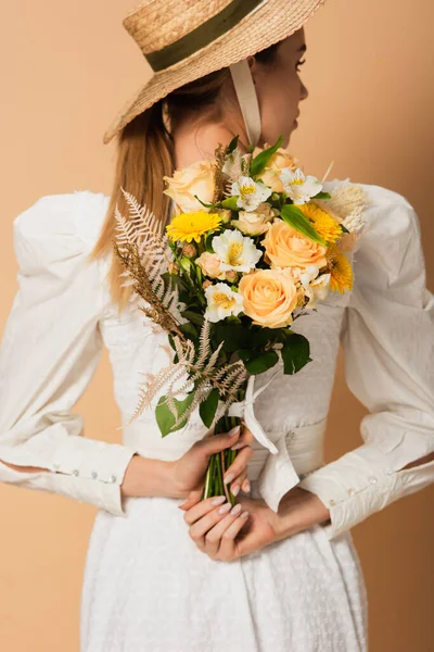 Young woman in straw hat holding bouquet of flowers behind back on beige — Stock Photo