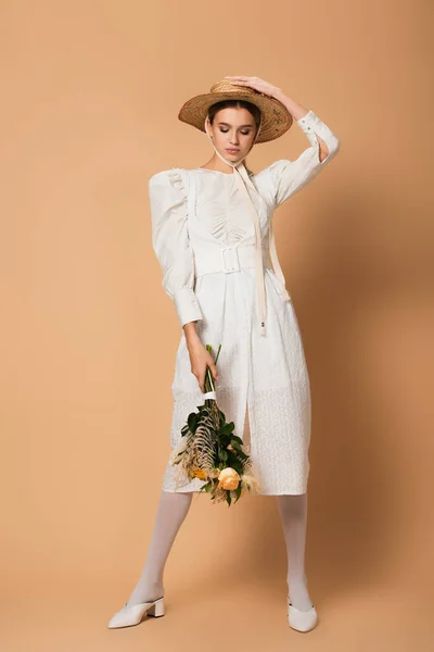 In voller Länge junge Frau in Kleid und Strohhut mit Blumenstrauß auf beige — Stockfoto