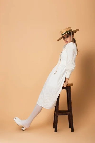 Full length of young woman in white dress and straw hat leaning on stool on beige — Stock Photo