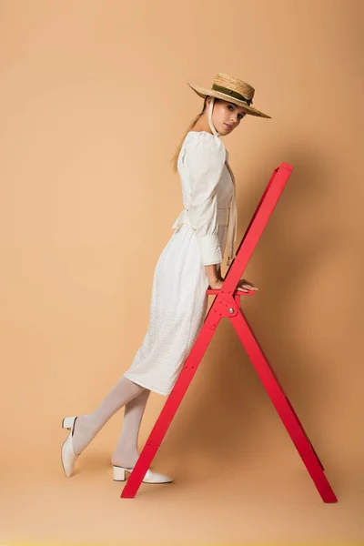 Full length of young woman in white dress and straw hat standing on red ladder on beige — Stock Photo