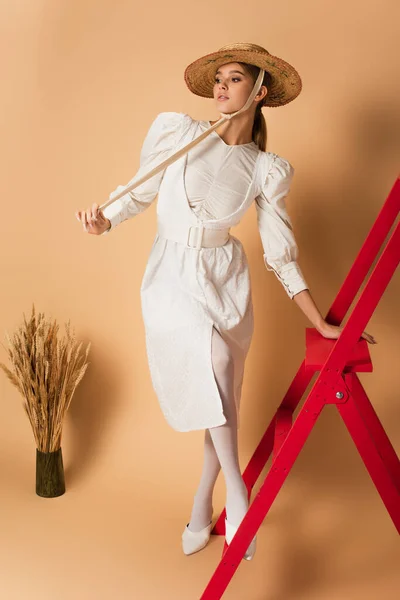 Полная длина молодой женщины в белом платье и соломенной шляпе позируя на красной лестнице возле вазы с шипами на бежевом — стоковое фото