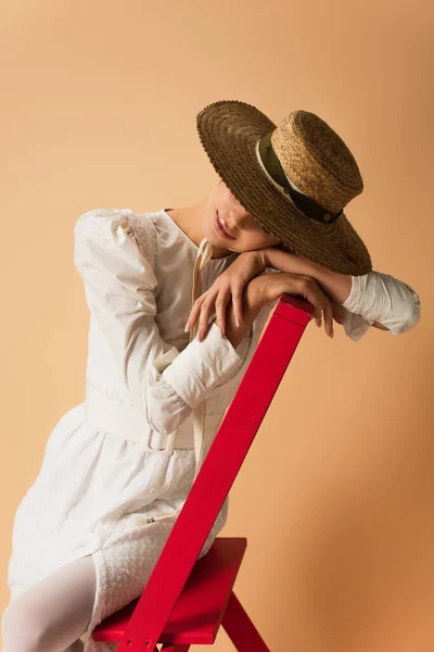 Mujer joven en vestido blanco y sombrero de paja apoyado en la escalera roja en beige - foto de stock