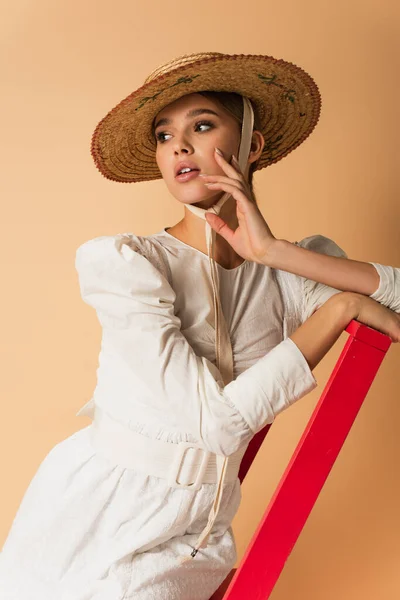 Giovane donna in abito e cappello di paglia in posa su scala rossa su beige — Foto stock