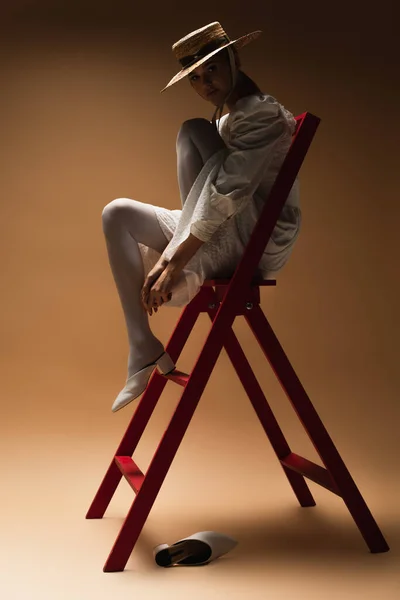 Giovane donna in abito bianco e cappello di paglia in posa su scala rossa in scuro su beige — Foto stock