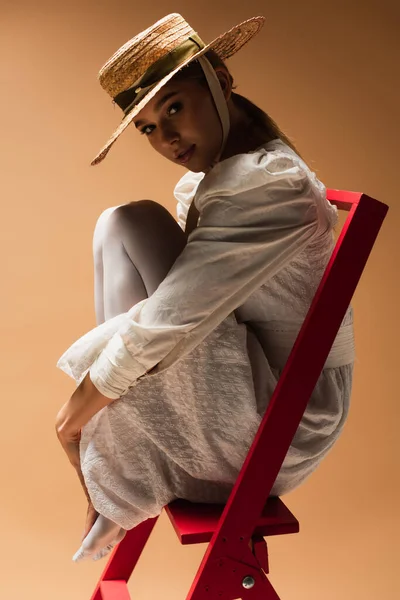 Jeune femme en robe blanche et chapeau de paille assis sur l'échelle rouge sur beige — Photo de stock