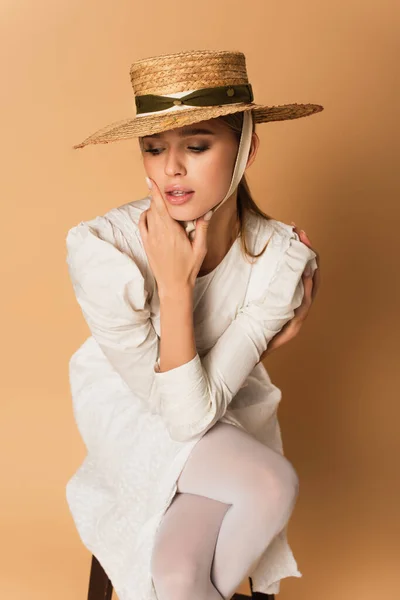 Pensive young woman in straw hat sitting on wooden chair on beige — Stock Photo