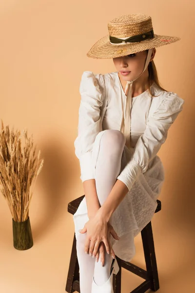 Mujer joven en sombrero de paja posando en silla de madera cerca de espiguillas de trigo en beige - foto de stock