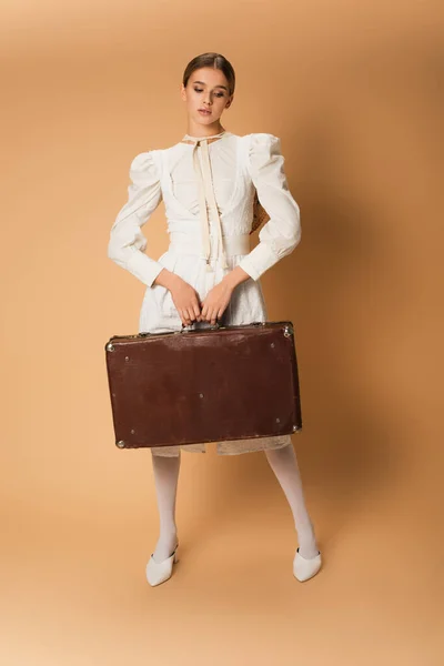 Full length of young woman in white dress holding suitcase on beige — Stock Photo