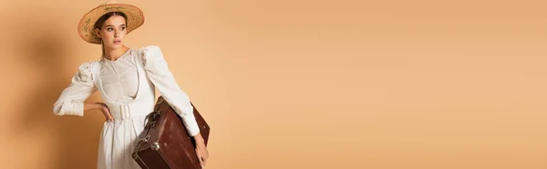 Jeune femme en robe blanche et chapeau de paille tenant une valise et posant à la main sur la hanche sur beige, bannière — Photo de stock