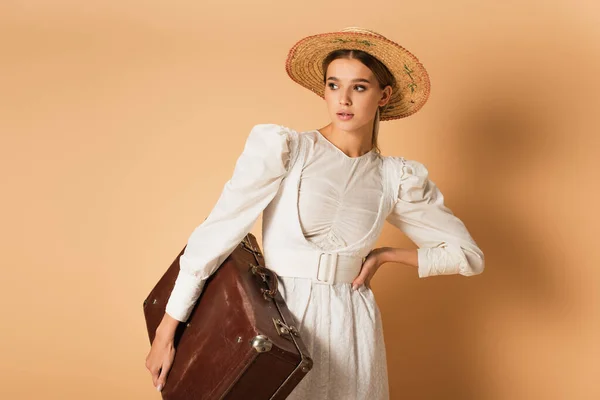 Jovem mulher em vestido branco e chapéu de palha segurando mala e posando com a mão no quadril no bege — Fotografia de Stock
