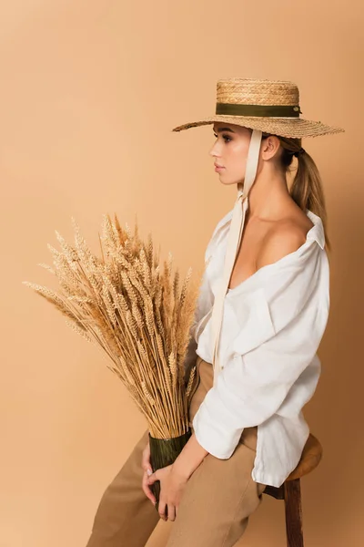 Side view of woman in white shirt and straw hat holding vase with wheat spikelets on beige — Stock Photo