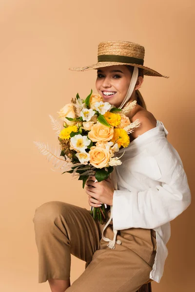 Sorrindo jovem mulher em camisa branca e chapéu de palha segurando buquê de flores no bege — Fotografia de Stock
