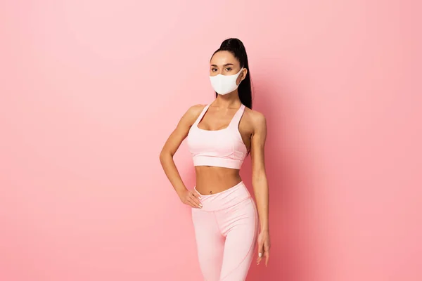 Young sportswoman in medical mask standing with hand on hip on pink — Stock Photo