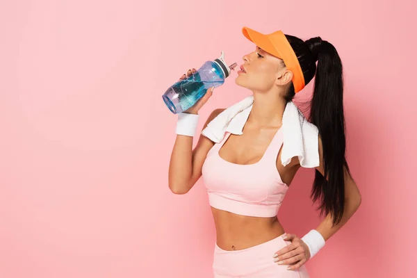 Vista lateral de una mujer deportiva con gorra de tenis sosteniendo una botella de deporte y agua potable en rosa - foto de stock