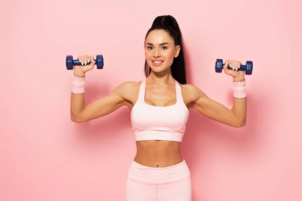 Радостная юная спортсменка, тренирующаяся с гантелями на розовом — стоковое фото