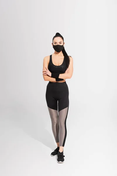 Jeune sportive en tenue de sport et masque de protection noir debout avec les bras croisés sur gris — Photo de stock