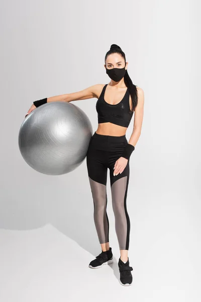 Young sportswoman in sportswear and black protective mask holding fitness ball on grey — Stock Photo