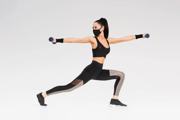 Full length of young sportswoman in black protective mask exercising with dumbbells on grey — Stock Photo