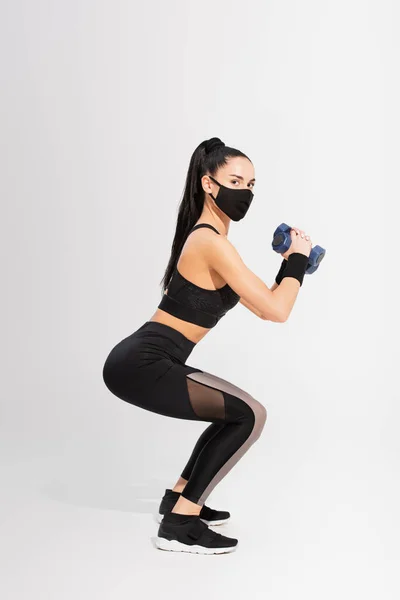 Full length of sportswoman in black protective mask exercising with dumbbells on grey — Stock Photo