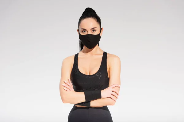 Jeune sportive en tenue de sport et masque de protection noir debout avec les bras croisés sur gris — Photo de stock