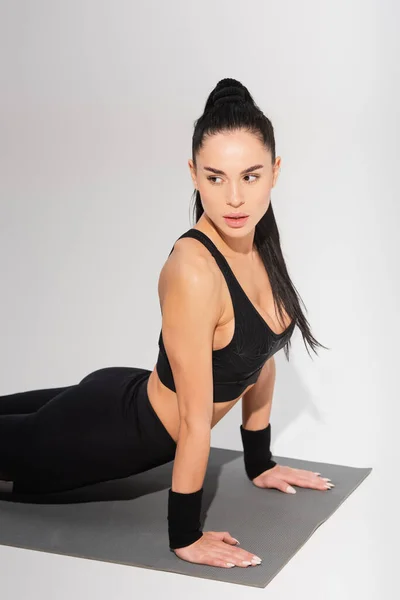 Jeune femme en sportswear noir faisant de l'exercice sur tapis de fitness sur gris — Photo de stock