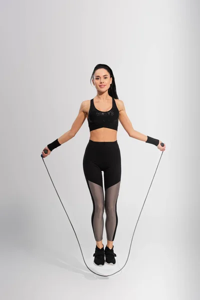 Full length of cheerful sportswoman exercising with skipping rope on grey — Stock Photo