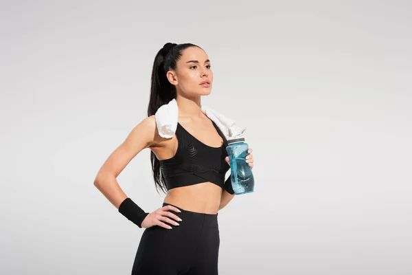 Молодая спортсменка с полотенцем на плечах держит спортивную бутылку, позируя рукой на бедре, изолированном на сером — стоковое фото