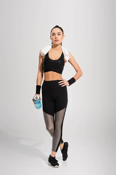 Full length of young sportswoman with towel on shoulders holding sports bottle while posing with hand on hip on grey — Stock Photo