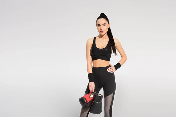Sportswoman holding boxing gloves while posing with hand on hip isolated on grey — Stock Photo
