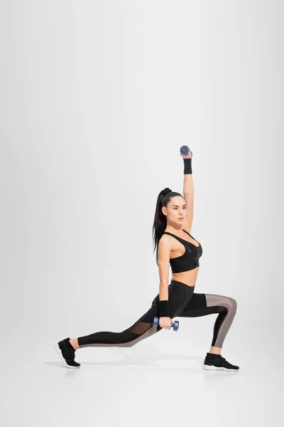 Full length of strong young sportswoman in sportswear working out with dumbbells on grey — Stock Photo