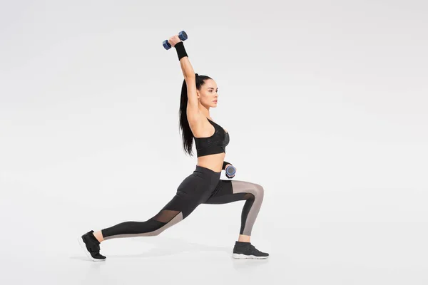 Full length of strong sportswoman in sportswear working out with dumbbells on grey — Stock Photo