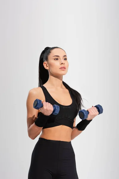 Сильная молодая спортсменка в спортивной одежде, тренирующаяся с гантелями, изолированными на сером — стоковое фото