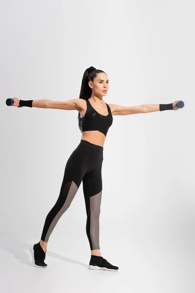 Full length of strong young sportswoman in sportswear exercising with dumbbells on grey — Stock Photo