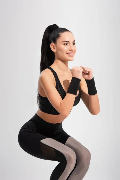 Young brunette sportswoman smiling while doing squat exercise isolated on grey — Stock Photo
