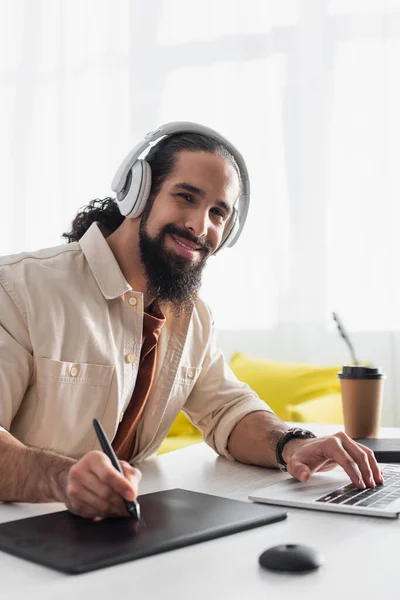 Lächelnder hispanischer Designer mit Kopfhörern, der am Laptop arbeitet und zu Hause Tablet zeichnet — Stockfoto